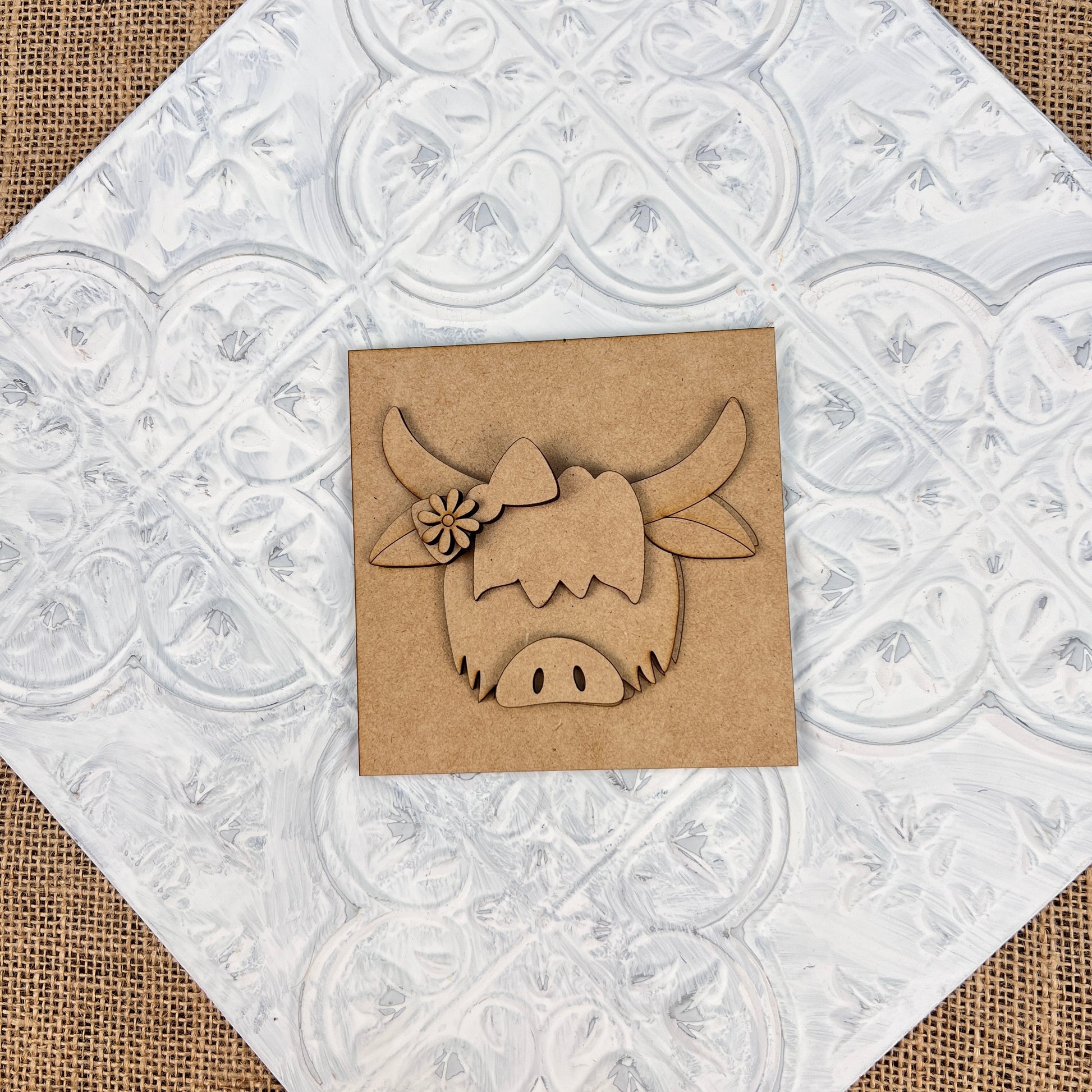 The "Farm Interchangeable Squares" by Janet's Craft Corner feature a charming woodcut of a highland cow adorned with a flower at the center, set against a textured white patterned tile background, perfect for embodying rustic farmhouse decor.