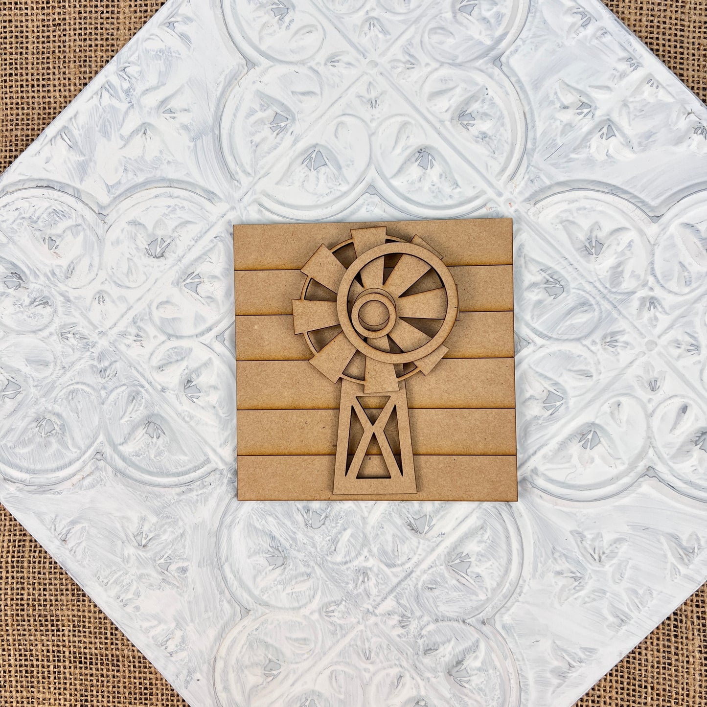 The *Farm Interchangeable Squares* by Janet's Craft Corner feature a wooden windmill design set against a square, textured metallic surface adorned with intricate floral patterns, capturing the essence of rustic farmhouse decor. The backdrop is crafted from burlap fabric, adding a touch of countryside charm.