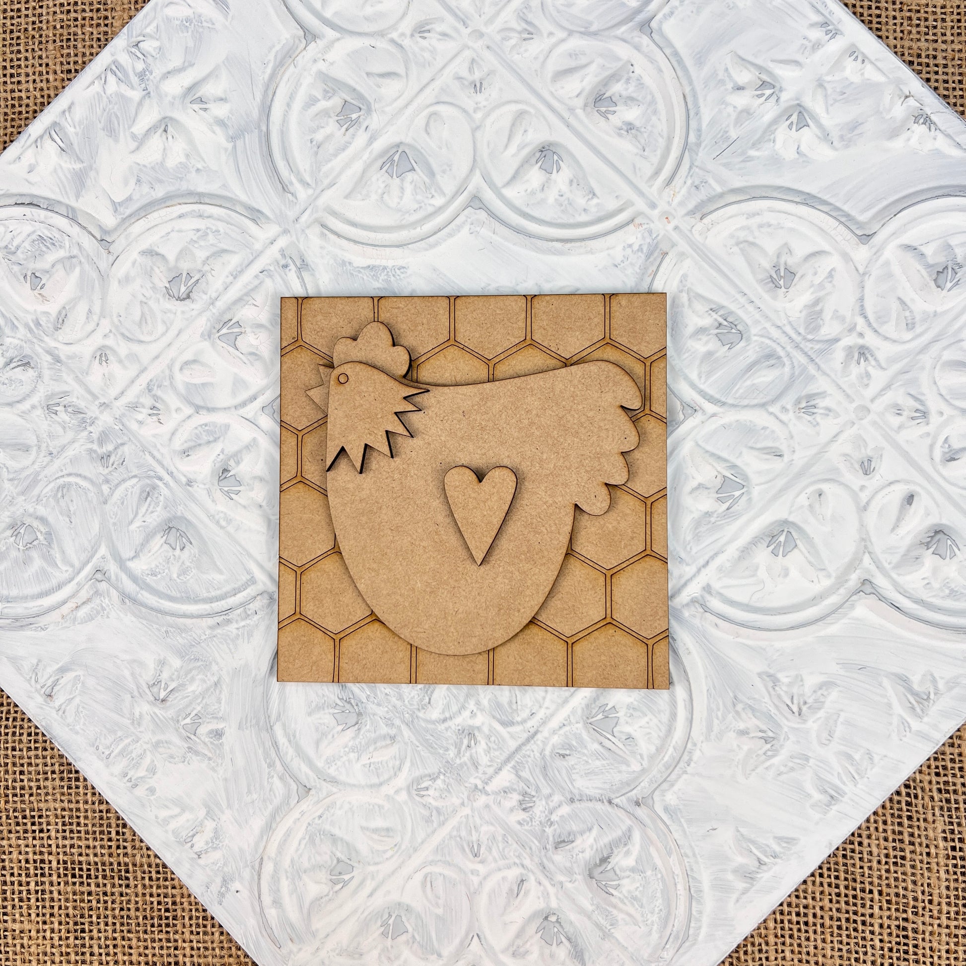 A charming piece of rustic farmhouse decor, the Farm Interchangeable Squares by Janet's Craft Corner features a chicken cutout with a heart on its body set against a honeycomb-patterned background. The white, textured floral design on burlap evokes the essence of countryside paradise.