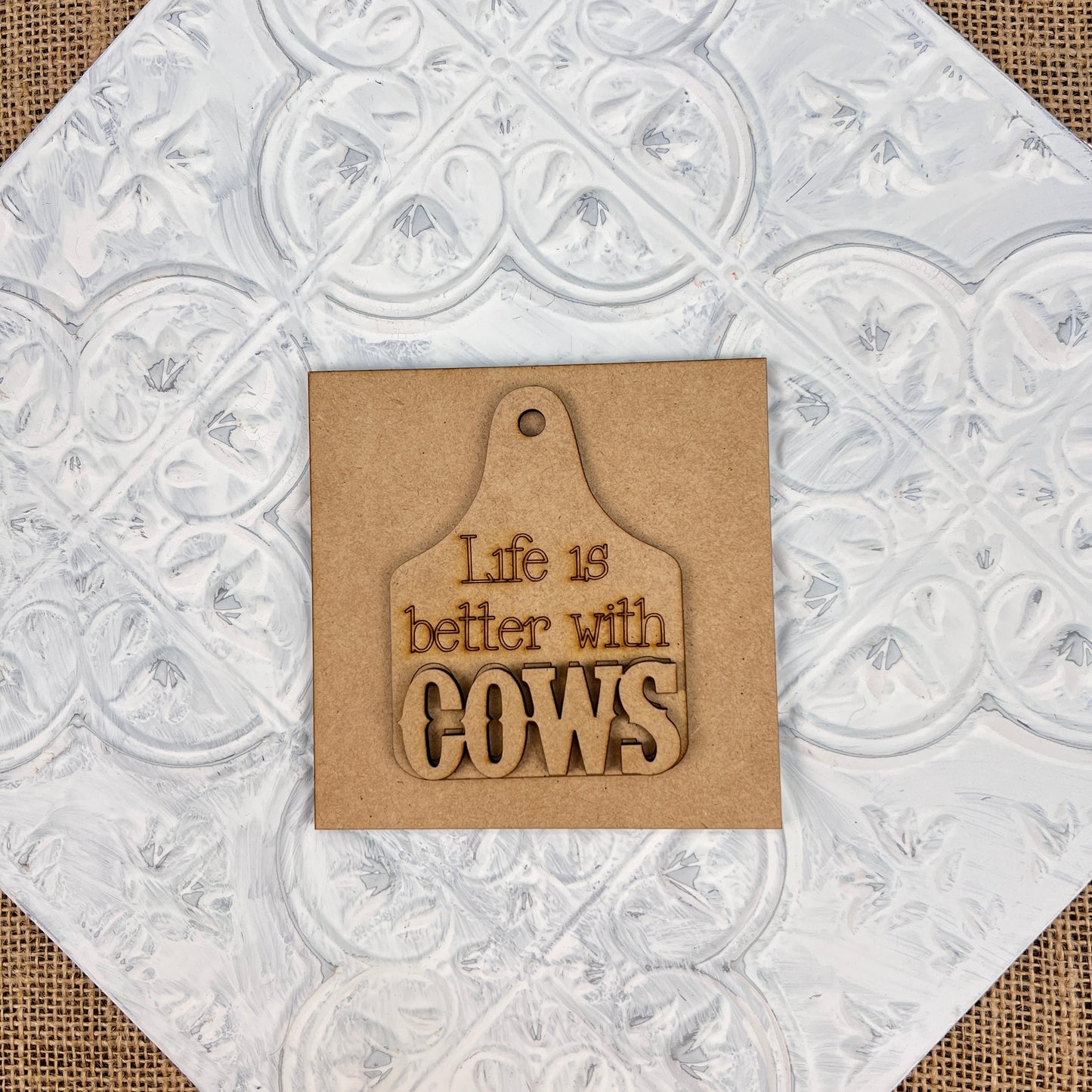 A quaint piece from Janet's Craft Corner, the Farm Interchangeable Squares feature a wooden plaque designed to evoke rustic farmhouse charm, set against a textured white background. Shaped like a cutting board and engraved with the phrase "Life is better with COWS," it makes for a delightful addition to any countryside haven.