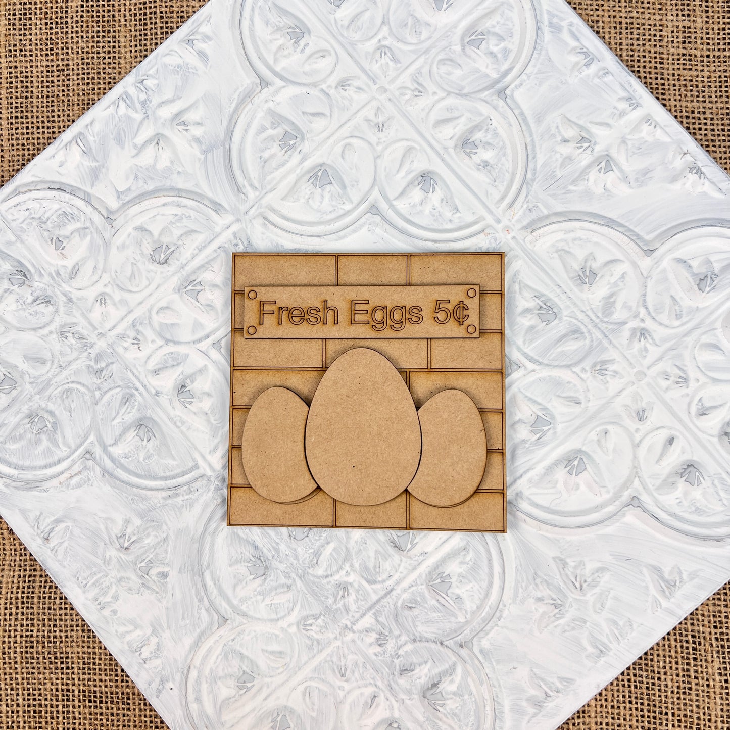 The "Farm Interchangeable Squares" by Janet's Craft Corner features a rustic wooden mini sign displaying "Fresh Eggs 5¢" alongside three egg shapes, all set against a textured white decorative background on burlap—making it a charming addition to your rustic farmhouse decor.