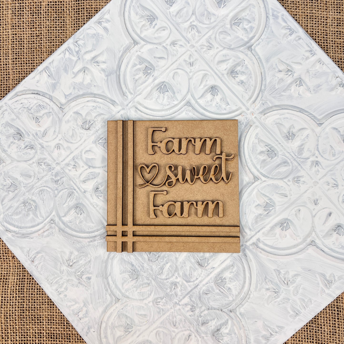 The "Farm Interchangeable Squares" by Janet's Craft Corner features a wooden square with "Farm sweet Farm" and a heart symbol, set against a white textured background with a vintage pattern. Placed on burlap fabric, this mini sign adds rustic farmhouse charm to any space, transforming it into a countryside paradise.