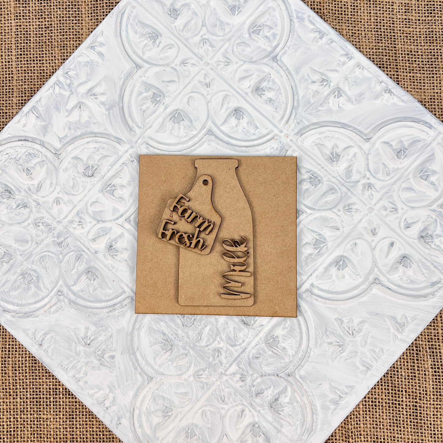 A square piece of brown cardboard from Janet's Craft Corner, featuring a milk bottle cutout and the phrase "Farm Fresh Milk," is set against a textured white and burlap background. This mini sign from the Farm Interchangeable Squares collection embodies rustic farmhouse decor, making it perfect for adding charm to tiered trays or any cozy corner.