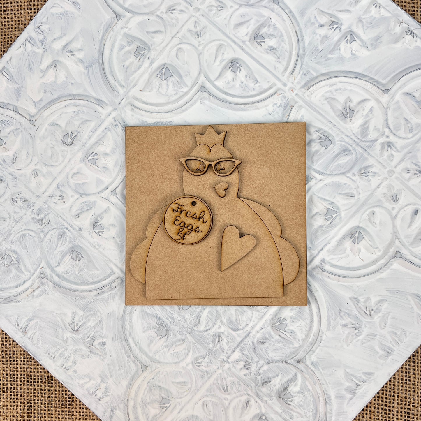 The Farm Interchangeable Squares by Janet's Craft Corner showcase a wooden cutout of a chicken wearing glasses, elegantly mounted on a brown square featuring a heart shape. The chicken is holding a small round sign that reads "Fresh Eggs." This delightful piece of farmhouse decor beautifully contrasts against the textured white background adorned with ornate patterns.