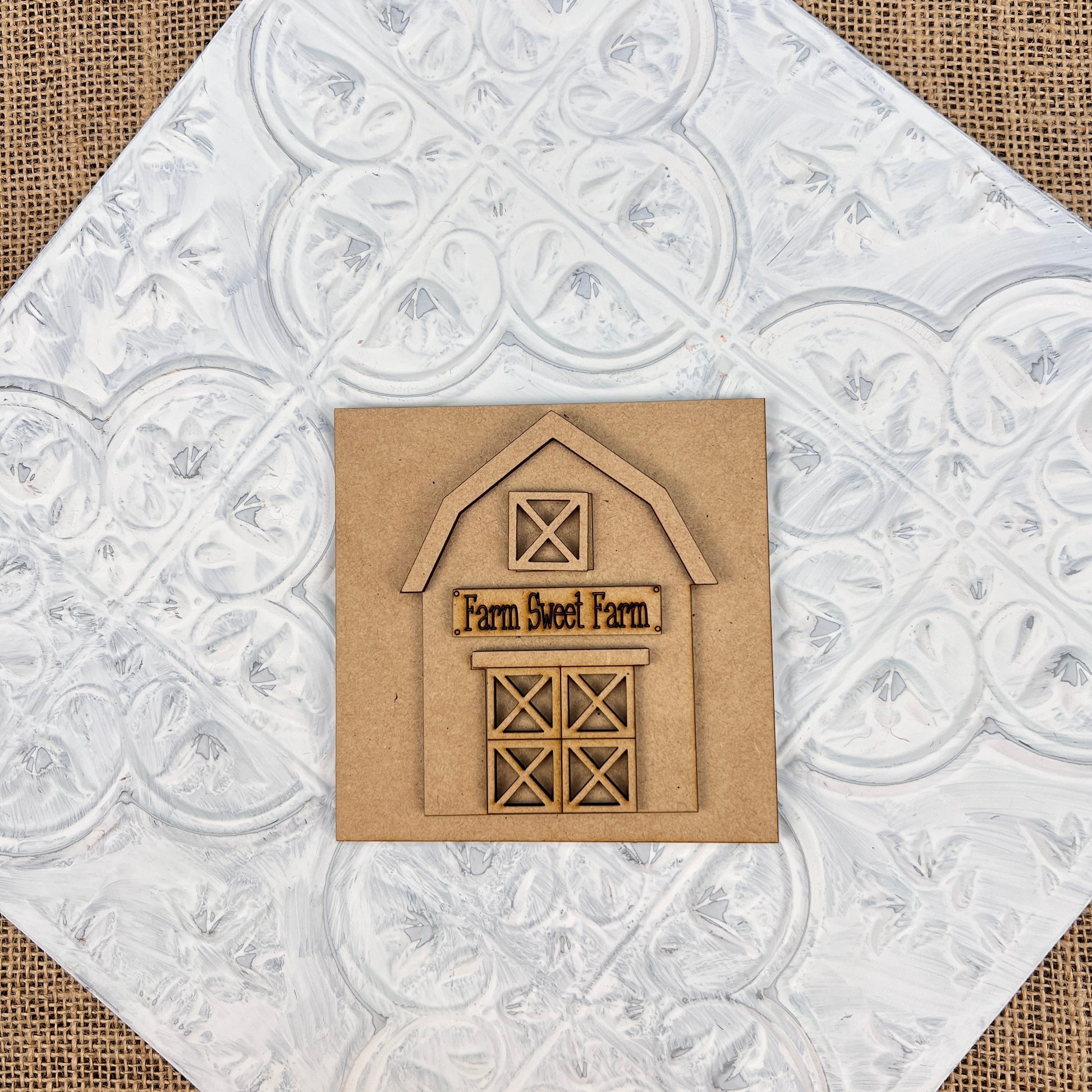 The "Farm Interchangeable Squares" from Janet's Craft Corner features a wooden barn cutout on a square cardboard piece, beautifully engraved with "Farm Sweet Farm." This delightful farmhouse decor is set against a textured white background adorned with circular patterns, reminiscent of vintage tin tiles, making it an ideal choice for infusing rustic charm into any countryside decoration.