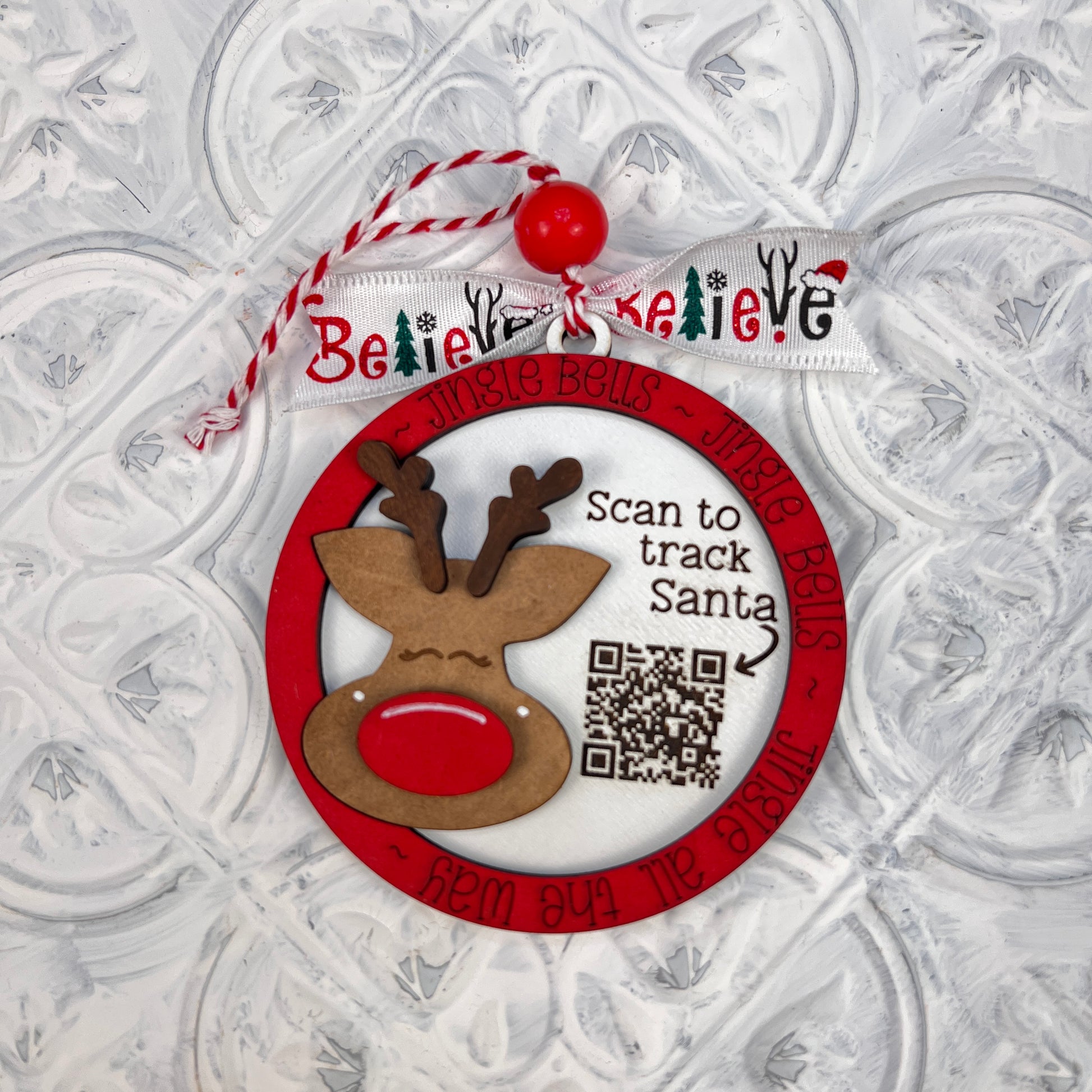 Product Description: The Santa Tracker Ornament from Janet's Craft Corner is a delightful, round DIY decoration kit perfect for festive holiday decor. This ornament showcases a charming reindeer face and includes a QR code labeled "Scan to track Santa." Adorned with the phrase "Jingle Bells" around its border, it also comes with a ribbon displaying the word "Believe" and is equipped with a red and white string finished with a red bead for hanging.