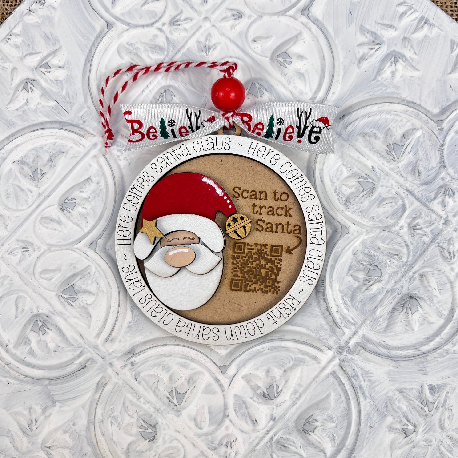 A round Christmas ornament from Janet's Craft Corner showcases Santa Claus, a QR code, and the phrase "Scan to track Santa" in the center. The perimeter features the repeated text "Here comes Santa Claus," making it perfect for holiday decor. It hangs with a red and white string adorned by a "Believe" ribbon, making it an ideal addition to your Santa Tracker Ornaments collection from the DIY Decoration Kit.