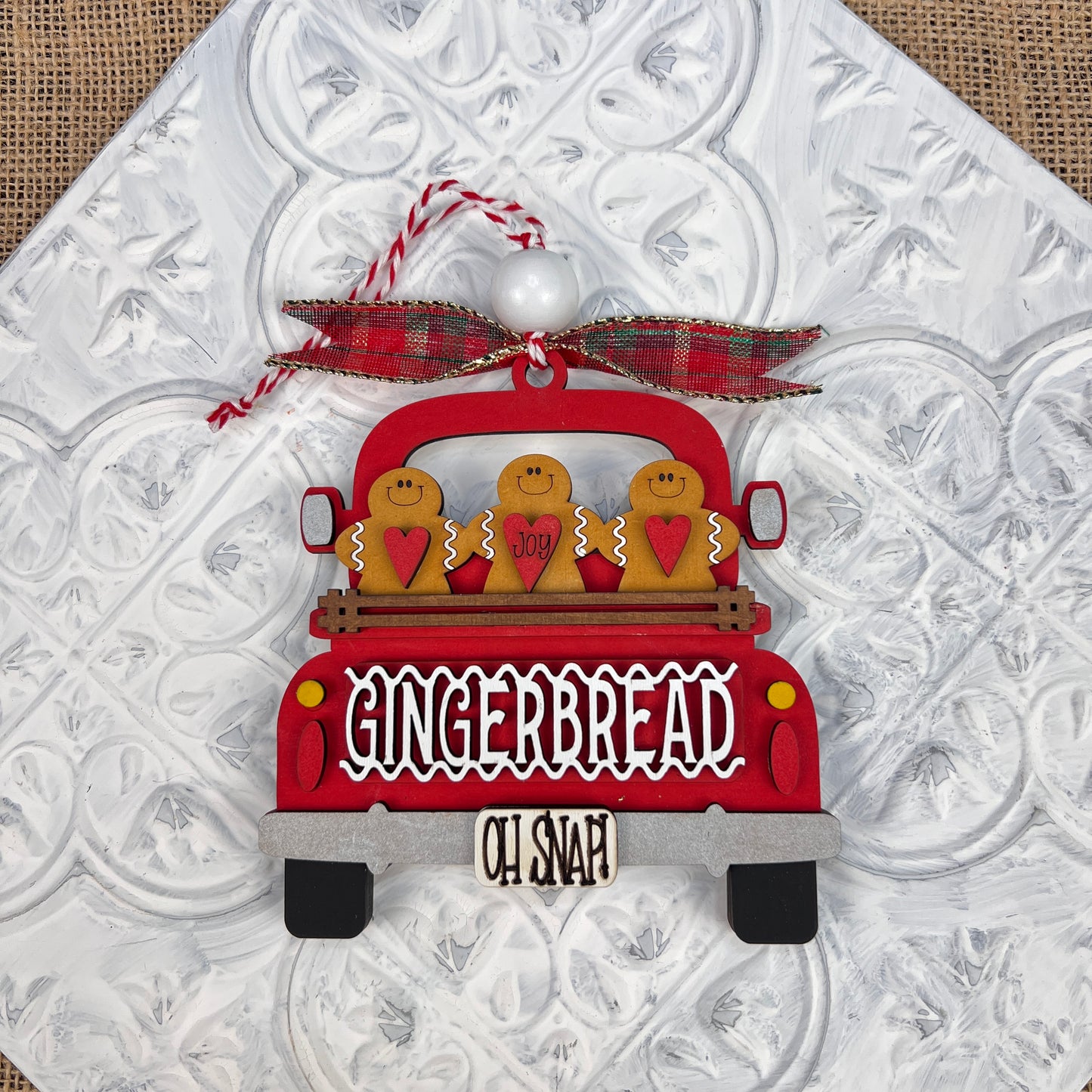 This delightful vintage-style piece, the Antique Truck Ornament - Painted by Janet's Craft Corner, features a red truck with "Gingerbread" written on it. It includes three gingerbread figures in the truck's bed and is adorned with a plaid bow and a tag that says "OH SNAP," all set against a textured white background.