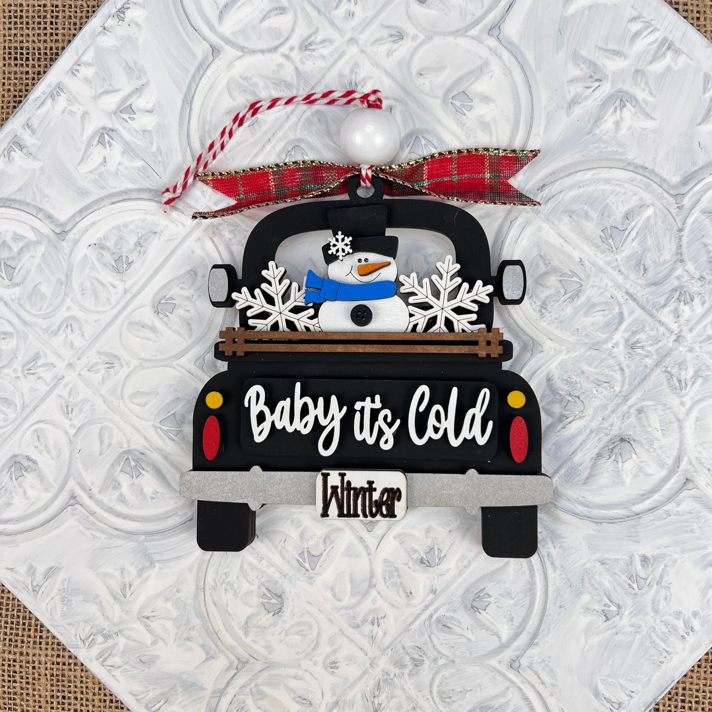 The Antique Truck Ornament - Painted from Janet's Craft Corner showcases a black antique truck featuring a snowman and snowflakes in its bed. It includes the phrases "Baby it's Cold" and "Winter" stylishly displayed, with a red plaid ribbon for easy hanging. Set against a patterned white background, this vintage-style piece is the perfect addition to your Christmas décor.