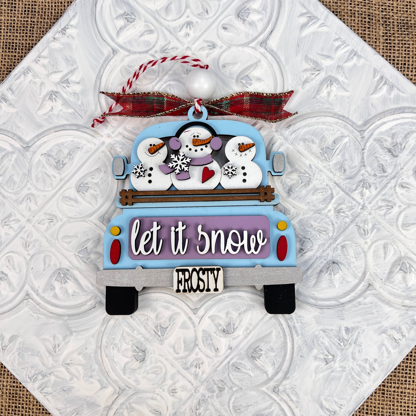 This nostalgic decoration from Janet's Craft Corner features the Antique Truck Ornament - Painted, showcasing a blue truck adorned with charming snowmen in hats and scarves. The phrase "Let it snow" is elegantly displayed on the body, while "Frosty" decorates the license plate. This hand-painted ornament is finished with a red plaid ribbon for an added touch of vintage charm.