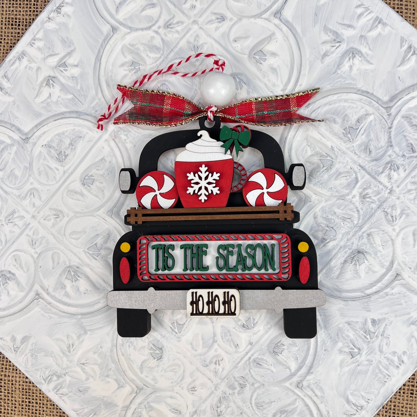 A vintage-inspired decoration from Janet's Craft Corner, the Antique Truck Ornament - Painted, showcases an antique truck carrying a mug with a snowflake design and two peppermint candies. The back of the truck reads "Tis the Season," while "Ho Ho Ho" graces the bumper, and it is topped with a festive plaid ribbon and red-and-white string.