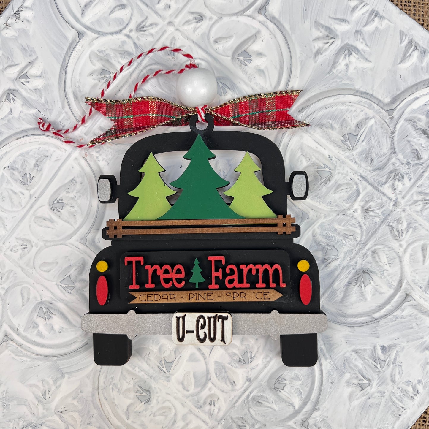 This charming addition to any holiday display is a vintage-style decoration from Janet's Craft Corner, featuring the "Antique Truck Ornament - Painted." It depicts an antique black truck carrying three green trees labeled "Tree Farm," adorned with a red plaid bow, and has a license plate that reads "U-CUT," all set against a textured white background.