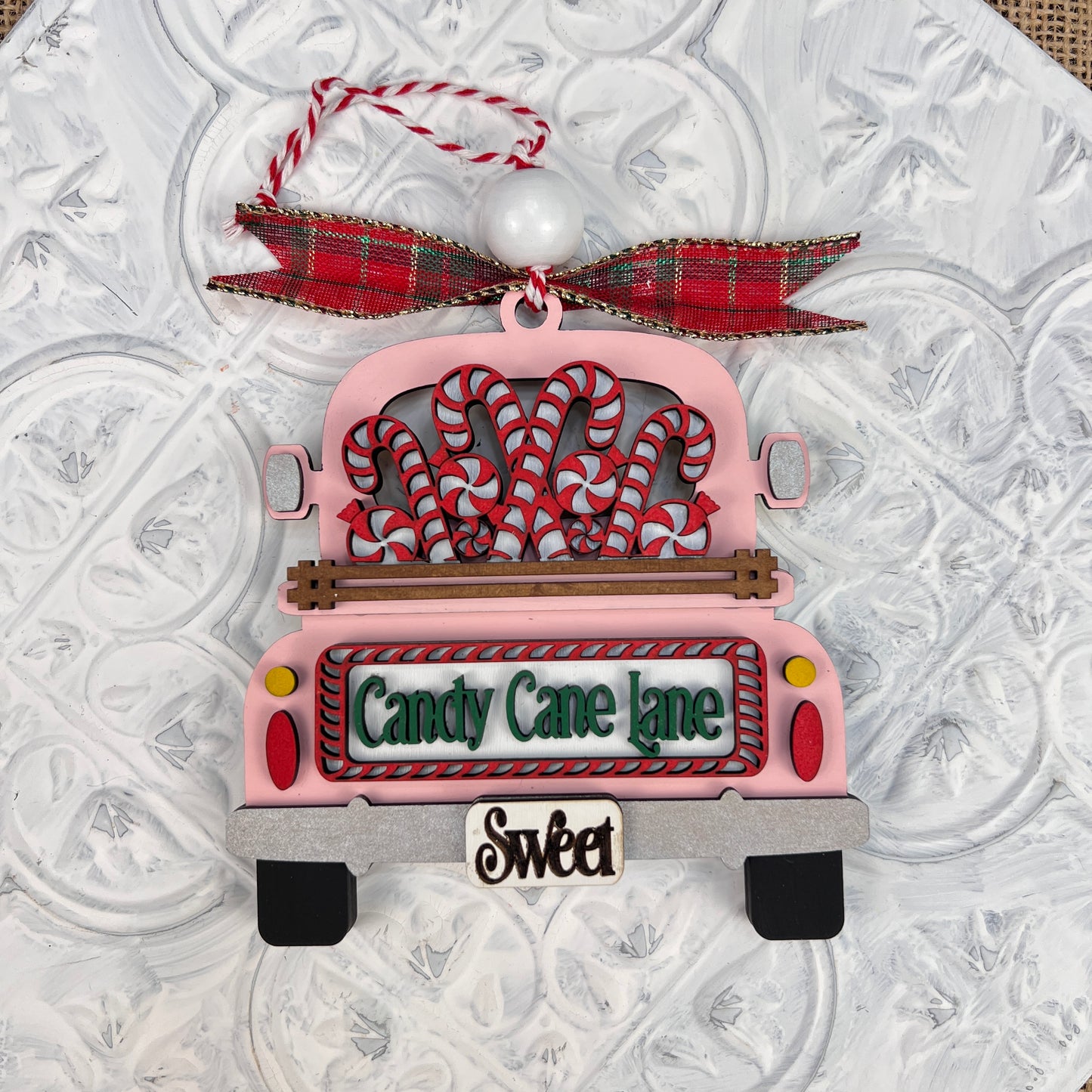 Bring charm to your holiday decor with the Antique Truck Ornament - Painted from Janet's Craft Corner. This vintage-style decoration features an antique truck filled with candy canes and adorned with a plaid ribbon on top. The license plate reads "Candy Cane Lane" with a small "Sweet" sign beneath, all beautifully set against a textured white surface.