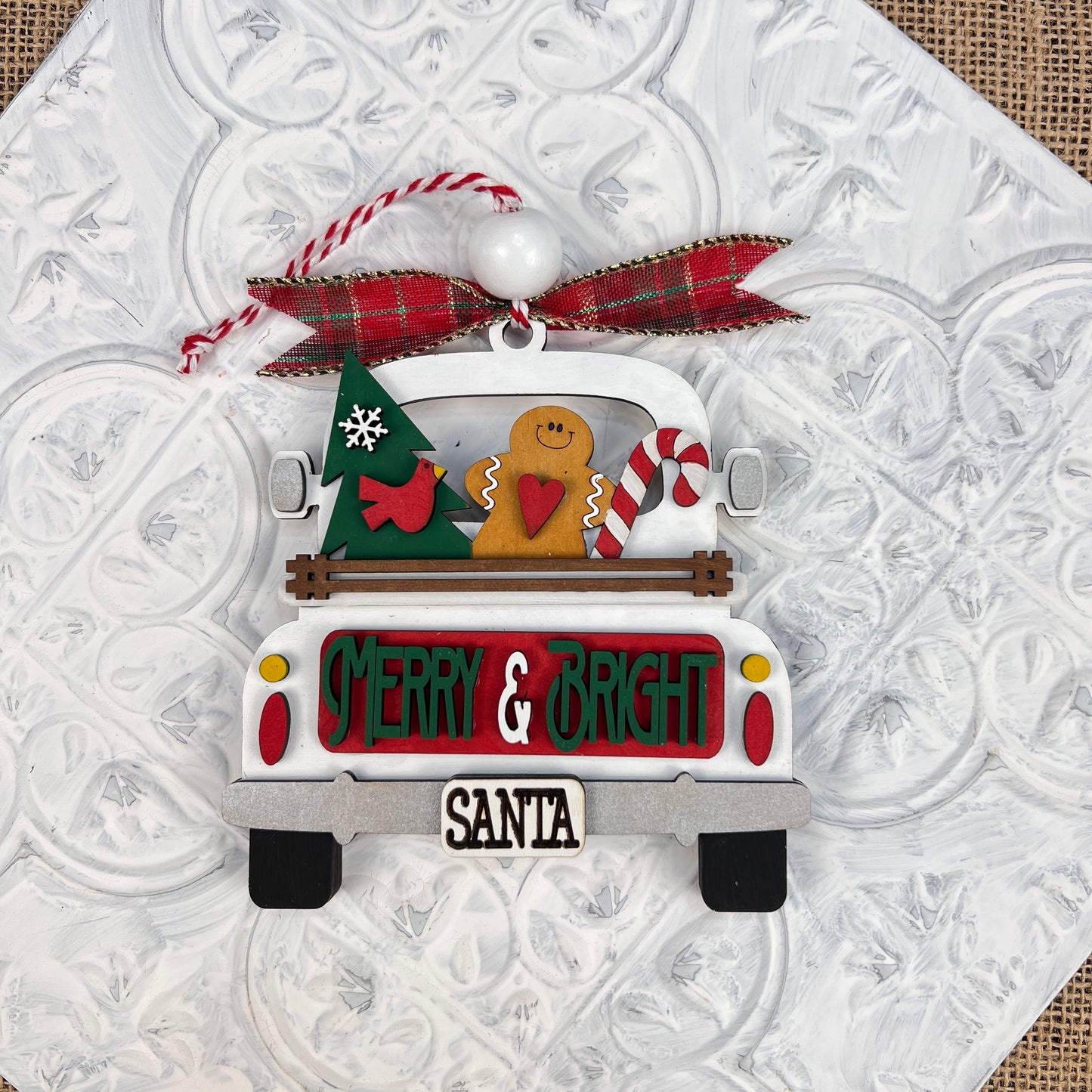 The "Antique Truck Ornament - Painted" from Janet's Craft Corner showcases a gingerbread man, tree, and candy cane in the back. Featuring the phrases "Merry & Bright" and "Santa," it is topped with a red plaid bow and bead for an extra charming touch to its vintage-style appearance.