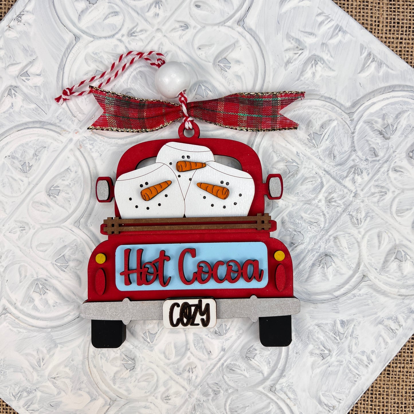 The "Antique Truck Ornament - Painted" by Janet's Craft Corner showcases a vintage-style red truck with cheerful snowmen and the words "Hot Cocoa." It is elegantly decorated with a festive red and green plaid ribbon and string, all set against a textured white background.