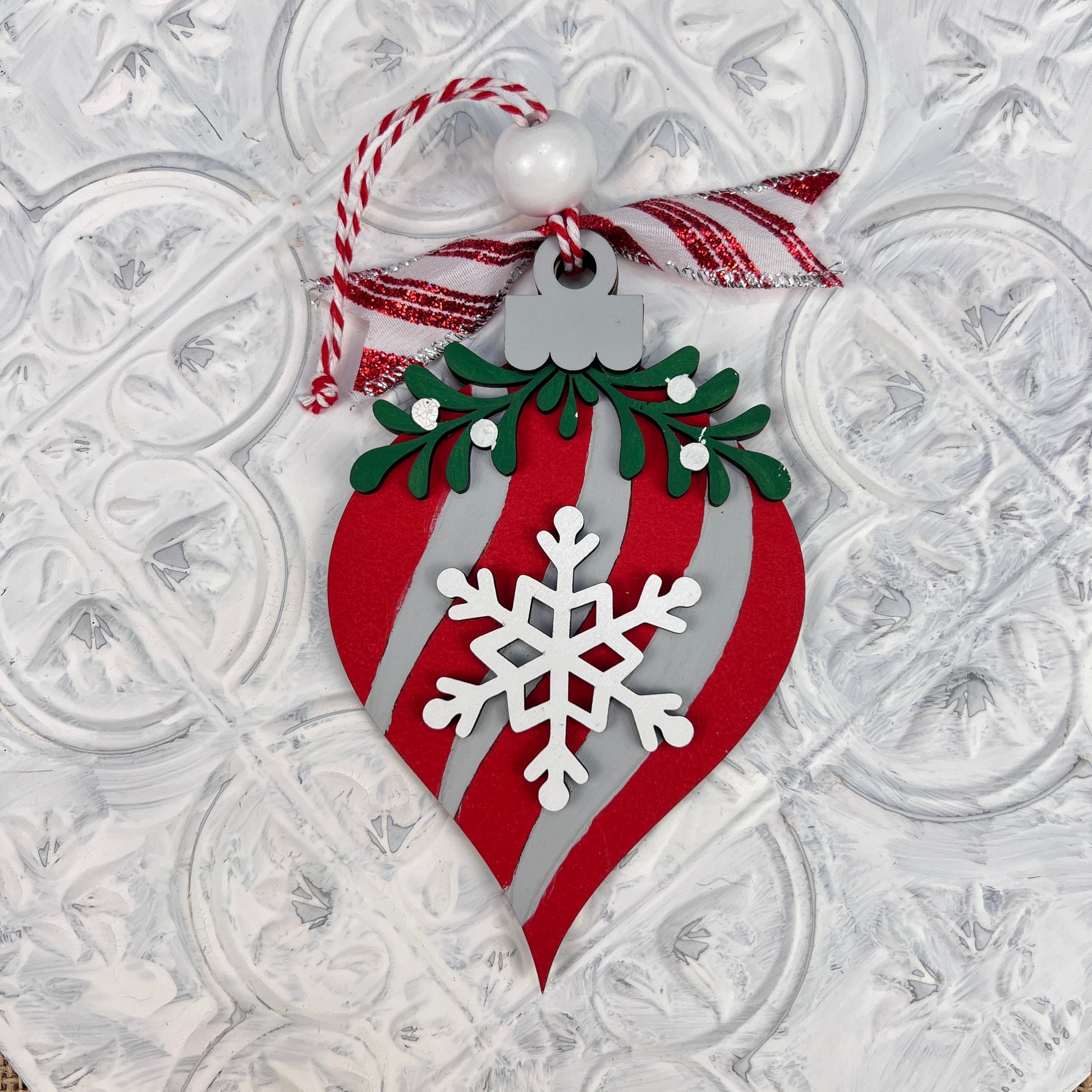 The Mistletoe Ornaments from Janet's Craft Corner feature a beautifully painted festive ornament with red and white stripes, showcasing a central snowflake design. It is adorned with mistletoe-like green foliage, complemented by a red and white ribbon and topped with a delicate white bead, all set against a textured white background.