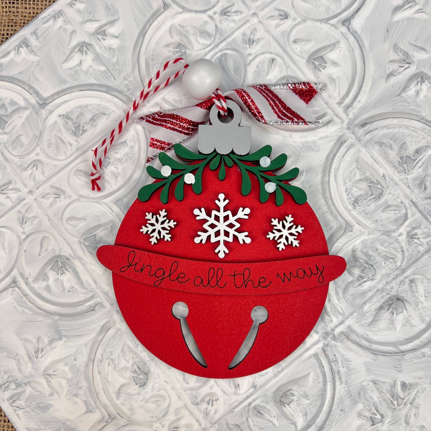 Introducing the Mistletoe Ornaments from Janet's Craft Corner, each individually hand-painted. This festive ornament is shaped like a red jingle bell adorned with green foliage and white snowflakes, featuring the phrase "Jingle all the way." It includes a charming white and red striped ribbon with a bead for hanging, set against an embossed white background.