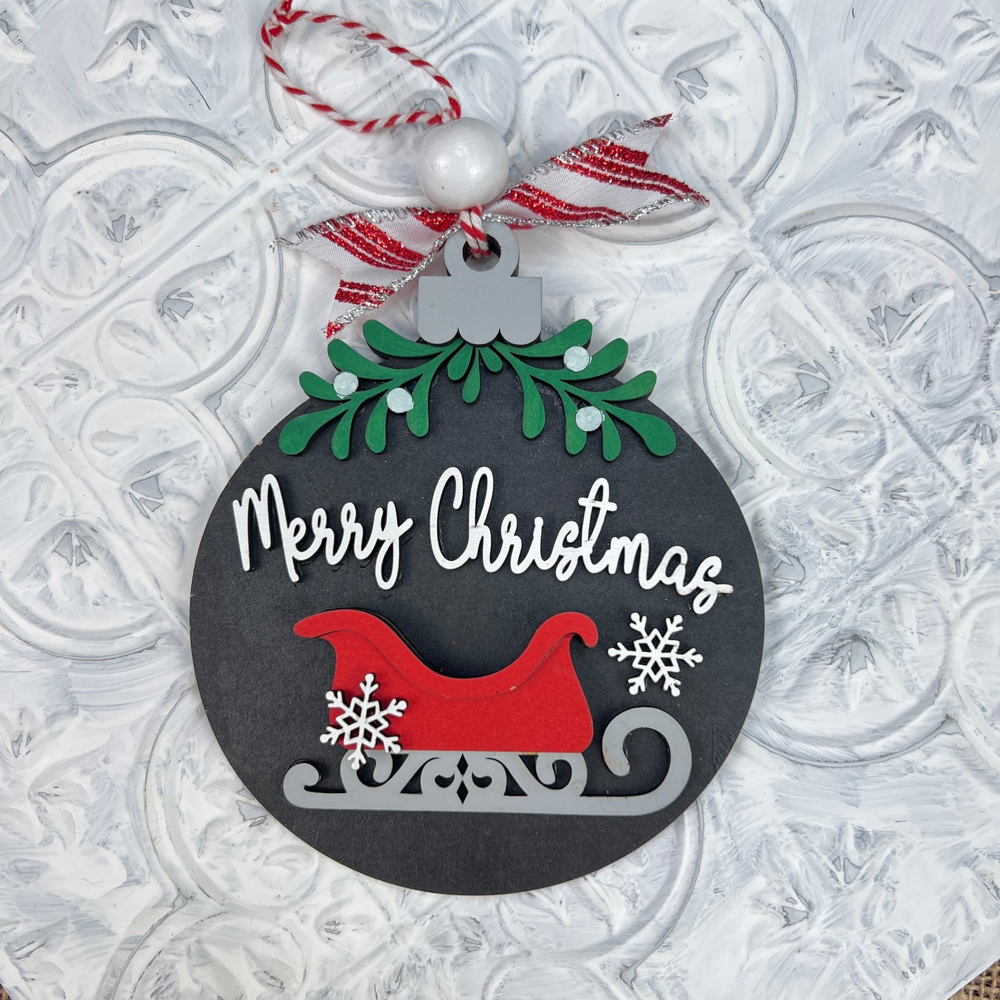 Experience holiday cheer with Janet's Craft Corner's beautifully hand-painted Mistletoe Ornaments. This round festive decoration features the text "Merry Christmas" in white, accompanied by a charming red and gray sleigh adorned with snowflakes. Topped with green holly and a red-and-white striped ribbon, it's the perfect addition to your festive collection.