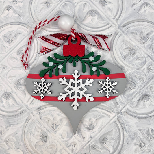 Introducing the "Mistletoe Ornaments - Individual Painted" by Janet's Craft Corner, a festive decoration showcasing a silver finish with three intricately hand-painted white snowflakes and green holly leaves. This ornament is elegantly accented with a red stripe and ribbon and hangs gracefully from a twisted red and white string against a textured white background.