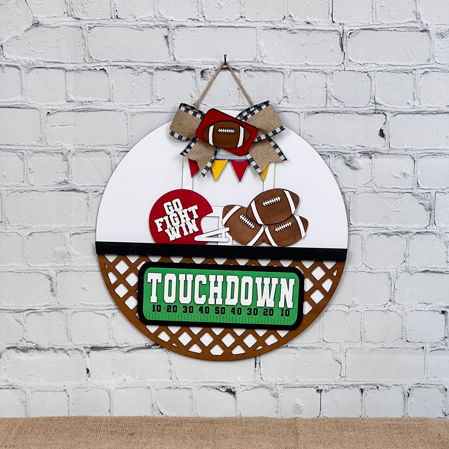 A round wooden sign from Janet's Craft Corner's Football Interchangeable Insert series is displayed on a white brick wall. It features a "Go Fight Win" banner with footballs, a ribbon, and a "Touchdown" scoreboard design complete with yard markers, making it an ideal hand-painted DIY craft kit for sports enthusiasts.