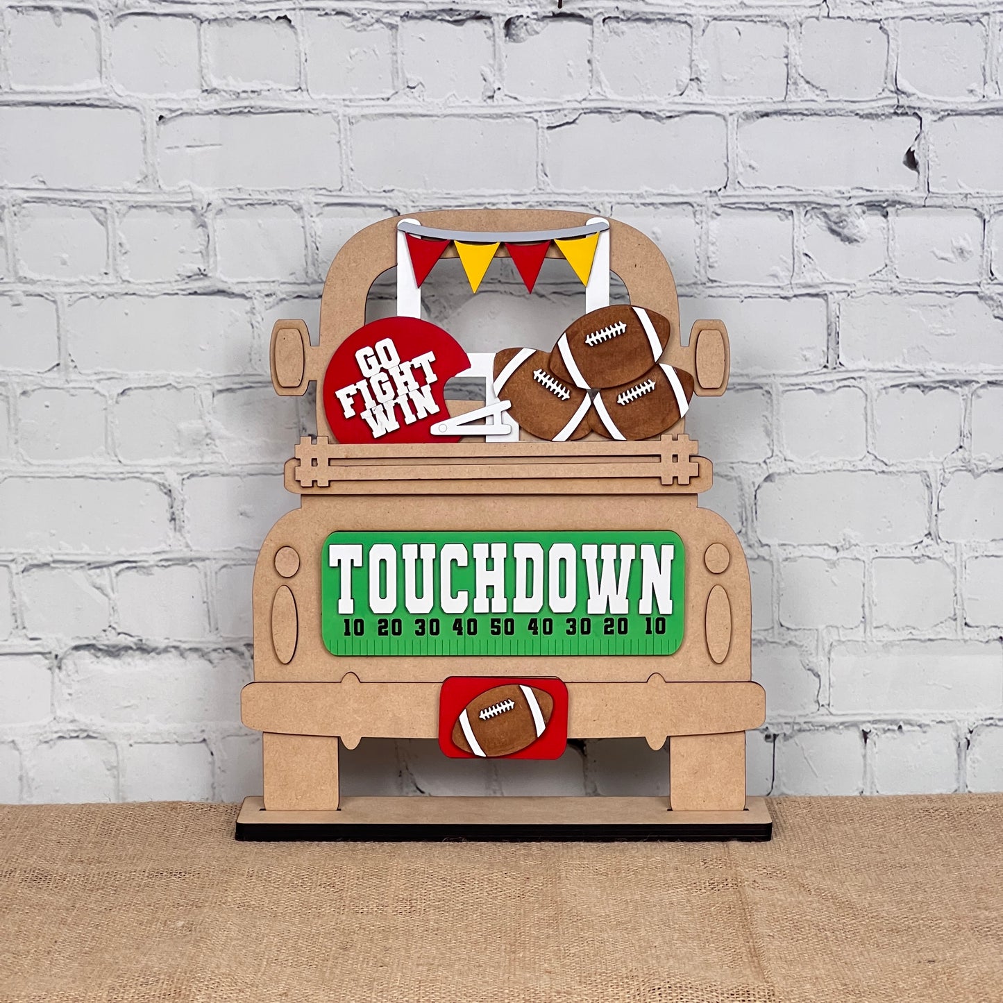 The Football Interchangeable Insert - Painted from Janet's Craft Corner showcases a football-themed decoration set against a white brick wall. It features a truck loaded with footballs, a "Touchdown" sign, and pennant flags. This versatile home decor piece is displayed on a burlap-covered surface, making it ideal for adding seasonal flair to your space.