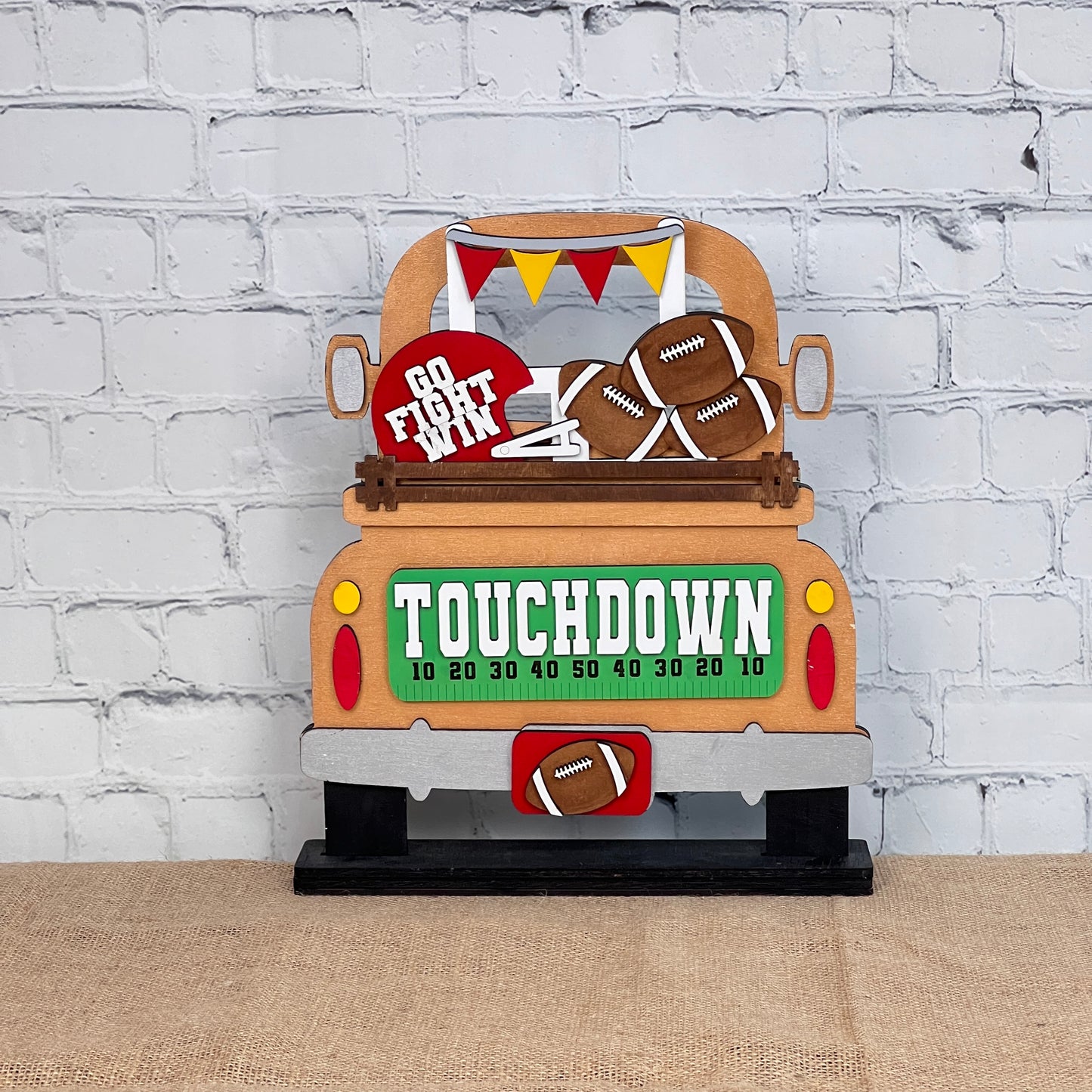 An interchangeable home decor piece featuring a decorative wooden cutout of a truck loaded with football-themed items. The truck displays "TOUCHDOWN" with yard line markers, alongside a "Go Fight Win" sign against a white brick wall backdrop. This is the Football Interchangeable Insert - Painted by Janet's Craft Corner.