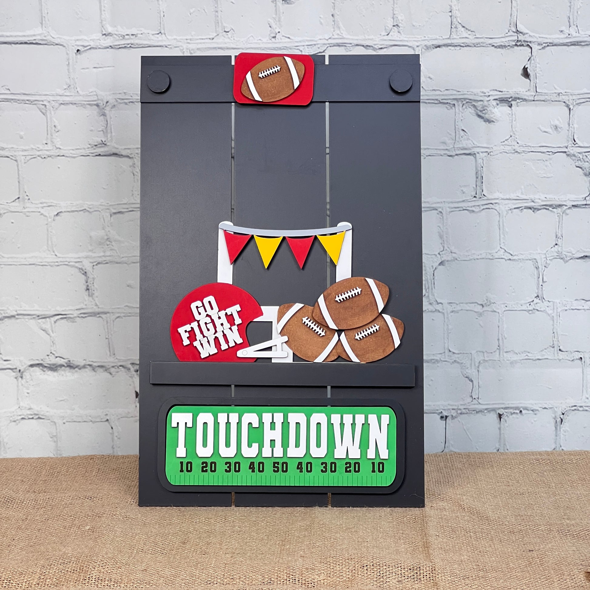 The Football Interchangeable Insert - Painted from Janet's Craft Corner is a DIY craft kit that includes hand-painted cutouts of footballs, a "Go Fight Win" sign, vibrant pennants, and a "Touchdown" strip marked with yard lines. This decorative board is set against a brick wall backdrop and placed on a burlap surface to enhance its charm.