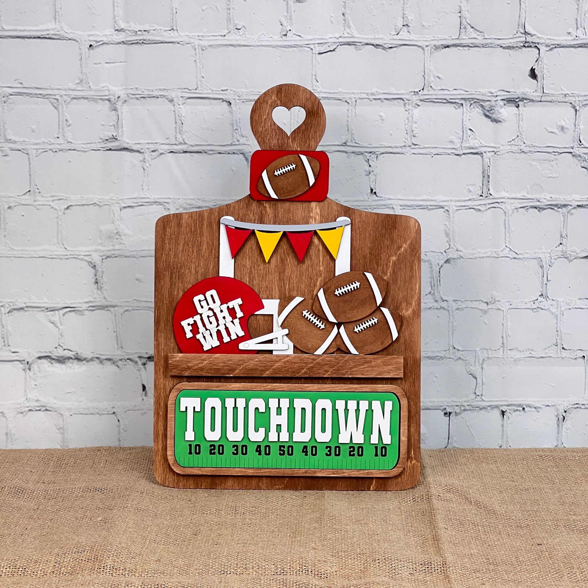 Introducing the Football Interchangeable Insert - Painted by Janet's Craft Corner: a wooden stand adorned with hand-painted football-themed decorations. This DIY craft kit features footballs, colorful banners, a mini scoreboard displaying "TOUCHDOWN," and a sign stating "GO FIGHT WIN." Set against a white brick wall, it's the perfect choice for interchangeable home decor.