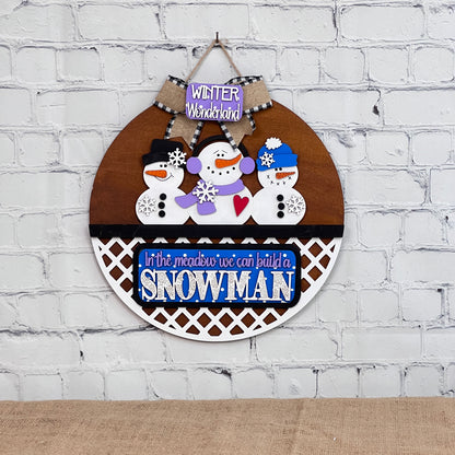 Janet's Craft Corner presents the "Snowman Interchangeable Insert," a round winter-themed wall decor kit featuring three snowmen with hats and scarves, titled "Winter Wonderland," and includes the phrase "In the meadow we can build a snowman." Perfect for DIY home decor against a white brick wall.
