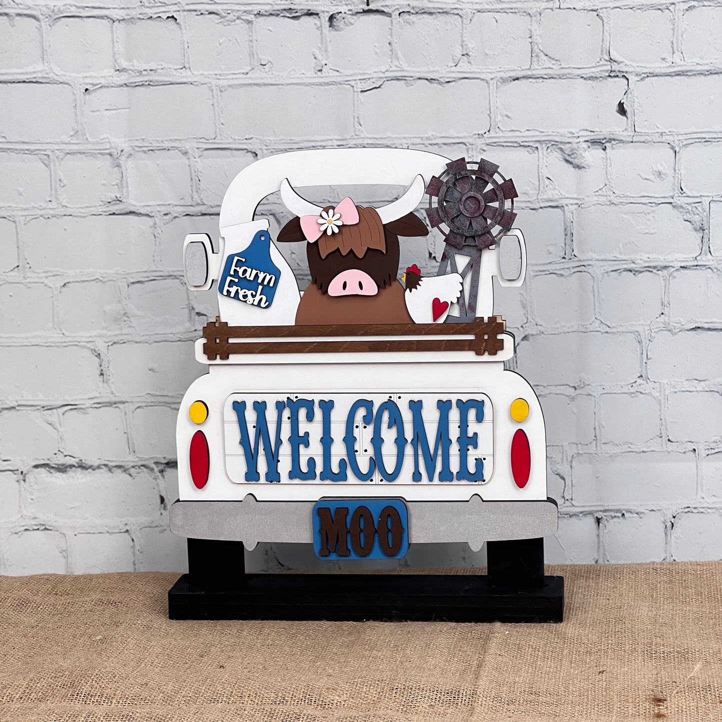 The Farm Welcome Interchangeable Insert by Janet's Craft Corner is a DIY home decor craft kit that includes three pieces: a decorative sign shaped like a truck with "WELCOME" and "MOO," featuring a Highland cow with flowers on its head holding a "Farm Fresh" sign against a brick wall backdrop. Perfect for enthusiasts.