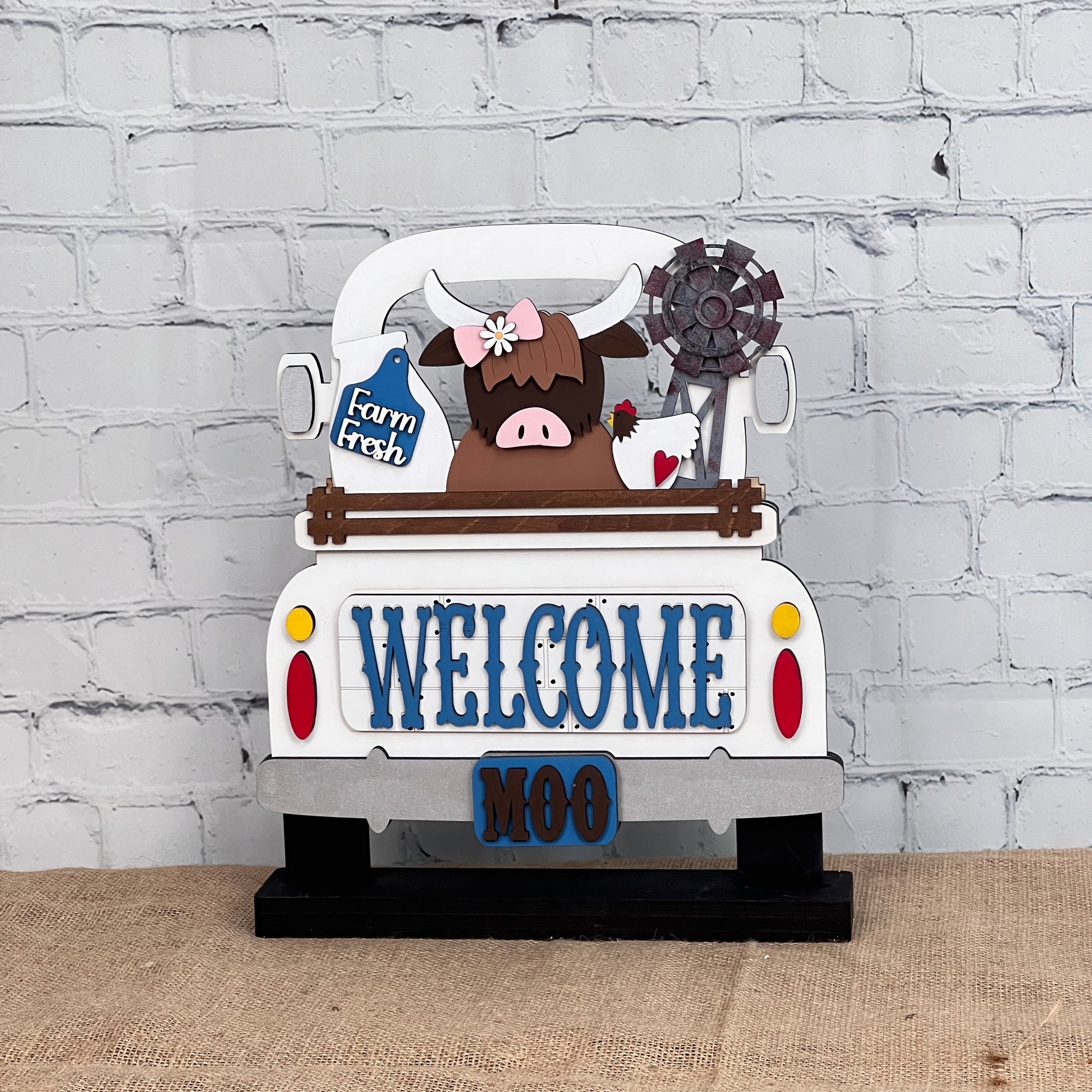 The Farm Welcome Interchangeable Insert by Janet's Craft Corner is a DIY home decor craft kit that includes three pieces: a decorative sign shaped like a truck with "WELCOME" and "MOO," featuring a Highland cow with flowers on its head holding a "Farm Fresh" sign against a brick wall backdrop. Perfect for enthusiasts.