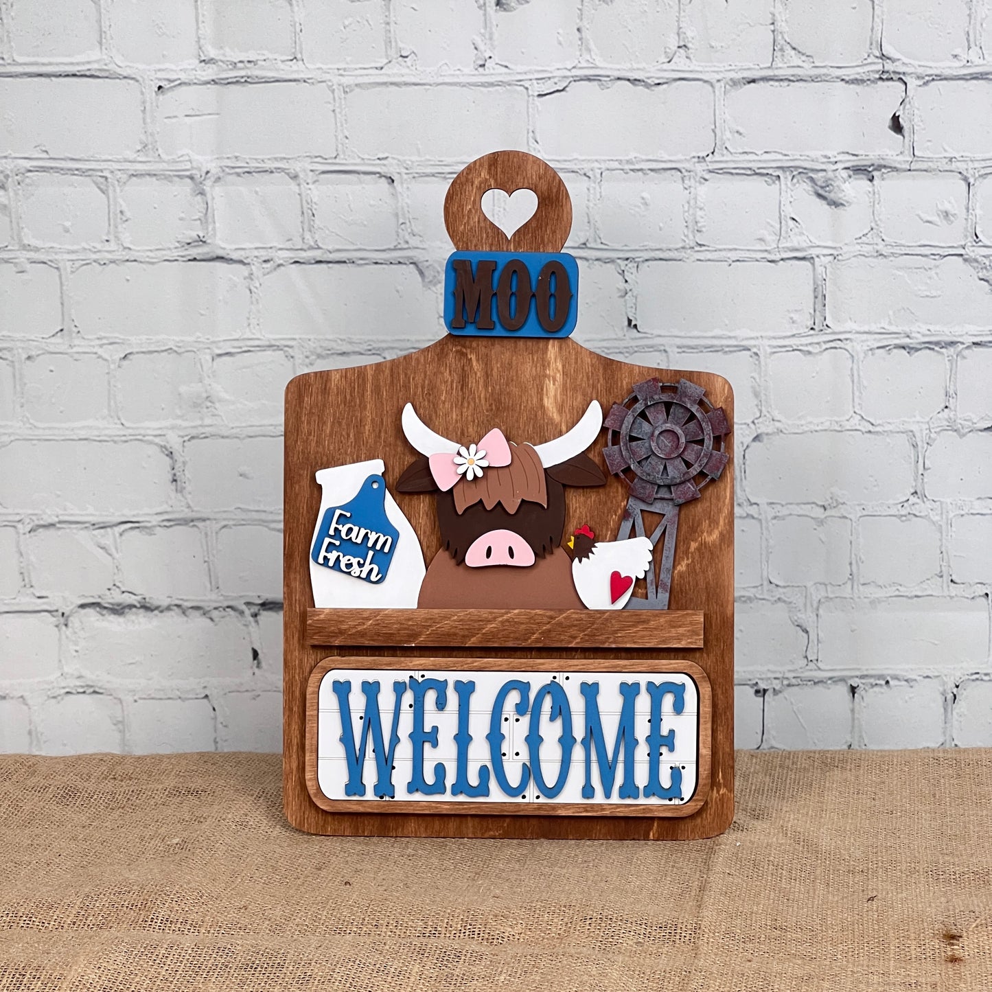 Janet's Craft Corner offers a DIY home decor kit: the Farm Welcome Interchangeable Insert, featuring a wooden board with "MOO" and "WELCOME" in blue, depicting a cartoon highland cow, milk bottle labeled "Farm Fresh," rusty windmill, and heart cutout. Perfect for burlap surfaces against white brick walls.