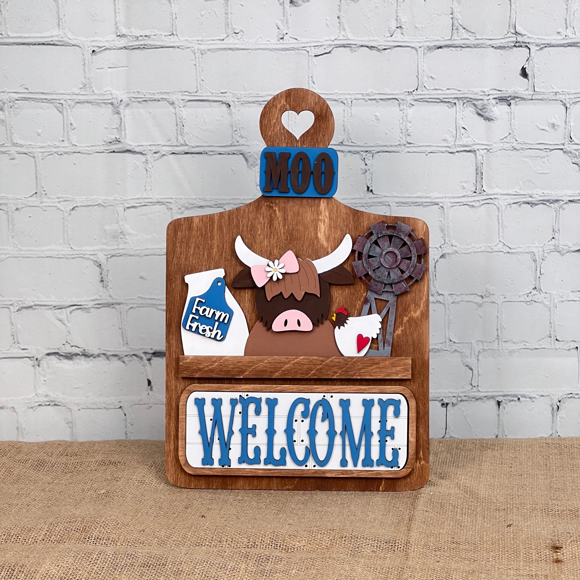 Janet's Craft Corner offers a DIY home decor kit: the Farm Welcome Interchangeable Insert, featuring a wooden board with "MOO" and "WELCOME" in blue, depicting a cartoon highland cow, milk bottle labeled "Farm Fresh," rusty windmill, and heart cutout. Perfect for burlap surfaces against white brick walls.