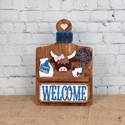 Janet's Craft Corner offers a DIY home decor kit: the Farm Welcome Interchangeable Insert, featuring a wooden board with "MOO" and "WELCOME" in blue, depicting a cartoon highland cow, milk bottle labeled "Farm Fresh," rusty windmill, and heart cutout. Perfect for burlap surfaces against white brick walls.
