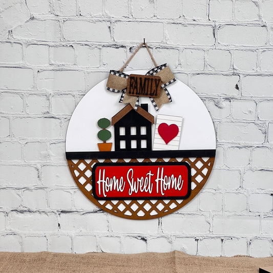 Against a white brick backdrop, a round wall sign displays a house, potted plant, and heart lattice. Topped with a "Family" bow, this Janet's Craft Corner DIY kit lets you create your own "Home Sweet Home" with interchangeable designs.