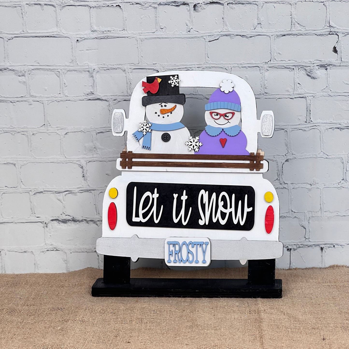 Janet's Craft Corner offers the "Let It Snow" DIY seasonal decor kit featuring an interchangeable insert with a pickup truck and snowman couple. The truck says "Let it snow" and has a "FROSTY" plate, adding winter cheer against its brick wall backdrop.