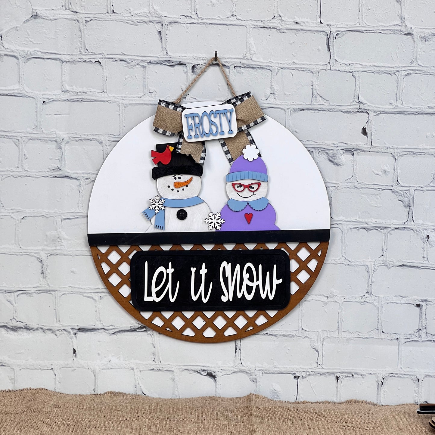 The Let It Snow Interchangeable Insert from Janet's Craft Corner features a snowman couple with hats, scarves, and glasses. One holds a broom under a "Frosty" sign with "Let it snow" beneath them on a white brick backdrop. Ideal for festive displays!.