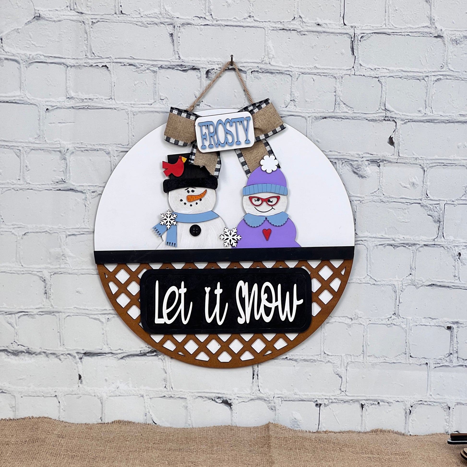 The Let It Snow Interchangeable Insert from Janet's Craft Corner features a snowman couple with hats, scarves, and glasses. One holds a broom under a "Frosty" sign with "Let it snow" beneath them on a white brick backdrop. Ideal for festive displays!.