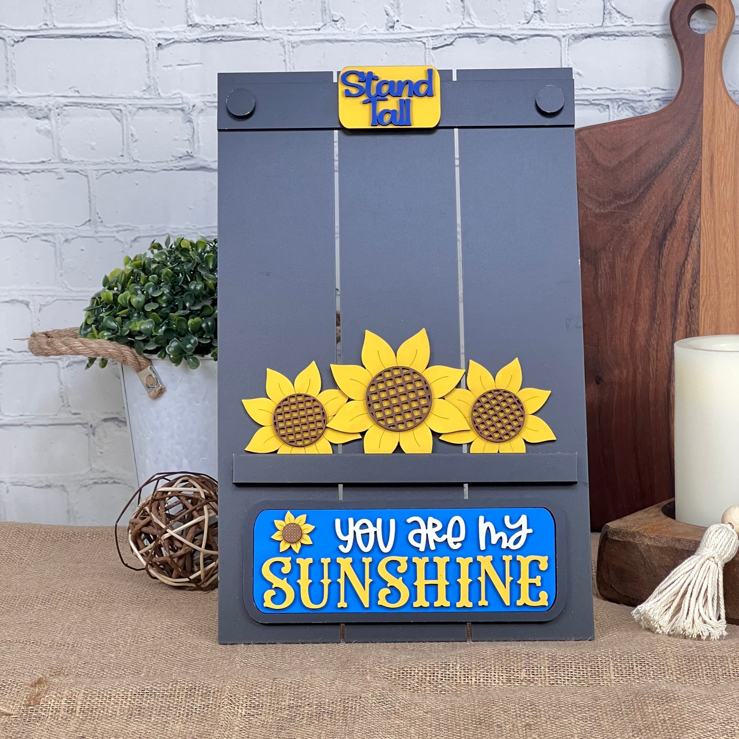 The Sunflower Interchangeable Insert from Janet's Craft Corner features a sunflower design with "Stand Tall" and "You Are My Sunshine" in blue and yellow. This decorative DIY home decor kit rests on burlap amidst plants and kitchen items.