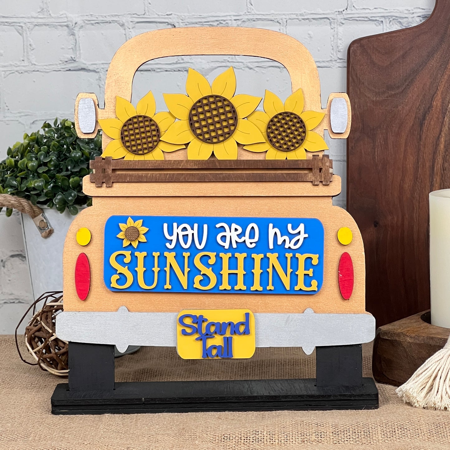 The Sunflower Interchangeable Insert from Janet's Craft Corner is a DIY home decor kit featuring a wooden truck cutout adorned with sunflowers and the phrase "You Are My Sunshine" in yellow and white, plus a small "Stand Tall" tag. Ideal for fans of interchangeable decorations, it complements any setting with plants, candles, or cutting boards.