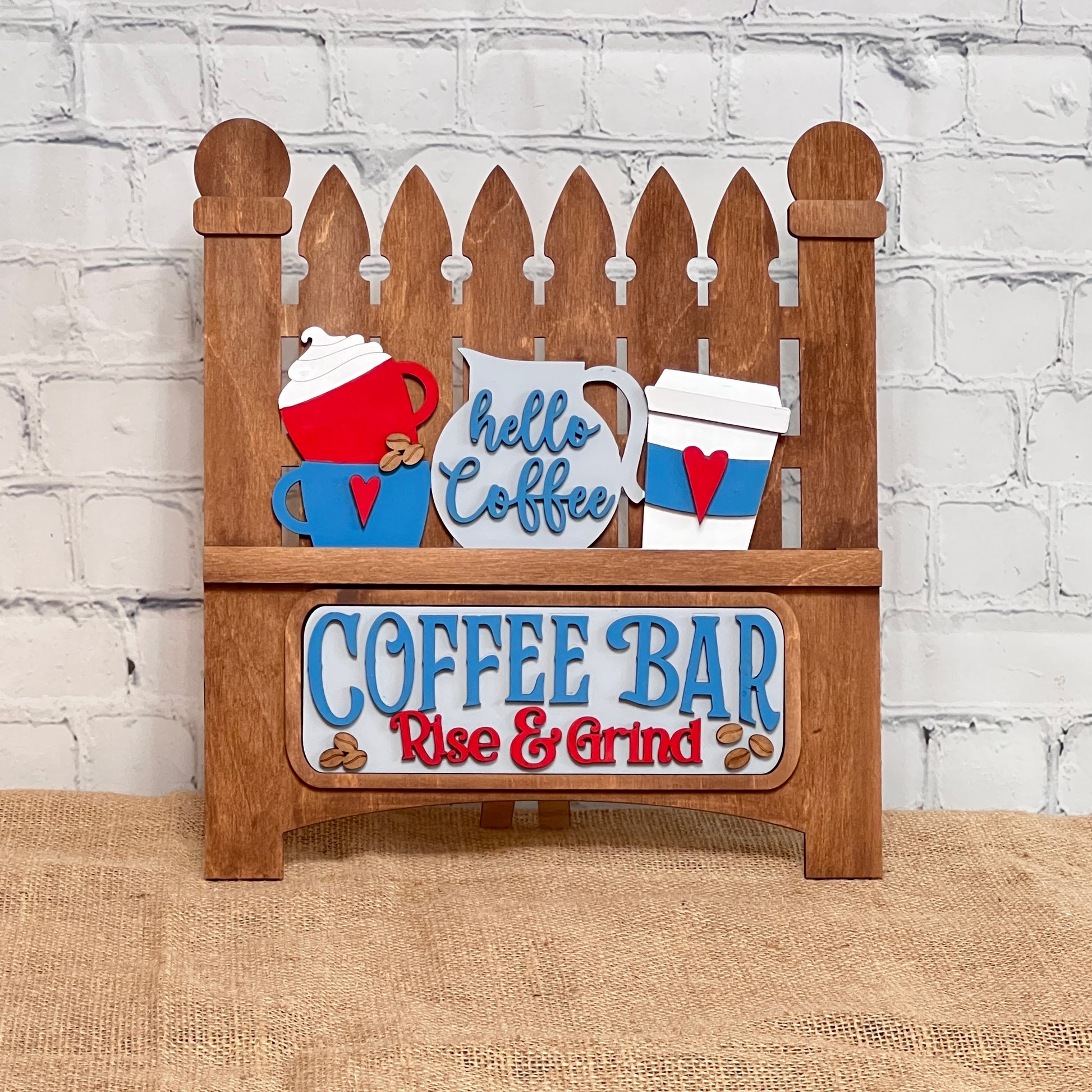 The Picket Fence with Insert from Janet's Craft Corner is a DIY interchangeable home décor kit featuring a charming hand-painted wooden design with colorful mugs and "hello coffee" above "COFFEE BAR Rise & Grind." Perfect for enhancing your home décor amidst a white brick and burlap setting.