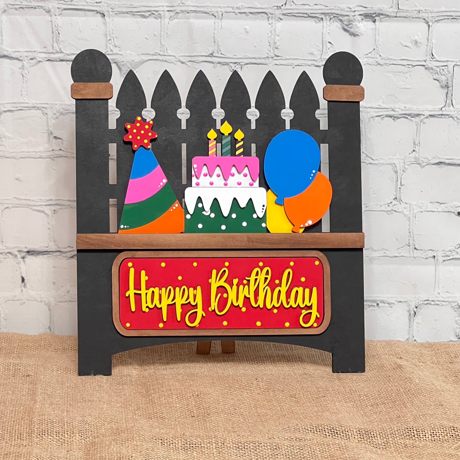 The Picket Fence with Insert by Janet's Craft Corner is a hand-painted wooden sign featuring a birthday cake, candles, party hat, and balloons against a white brick background. "Happy Birthday" is displayed on a red plaque at the bottom.