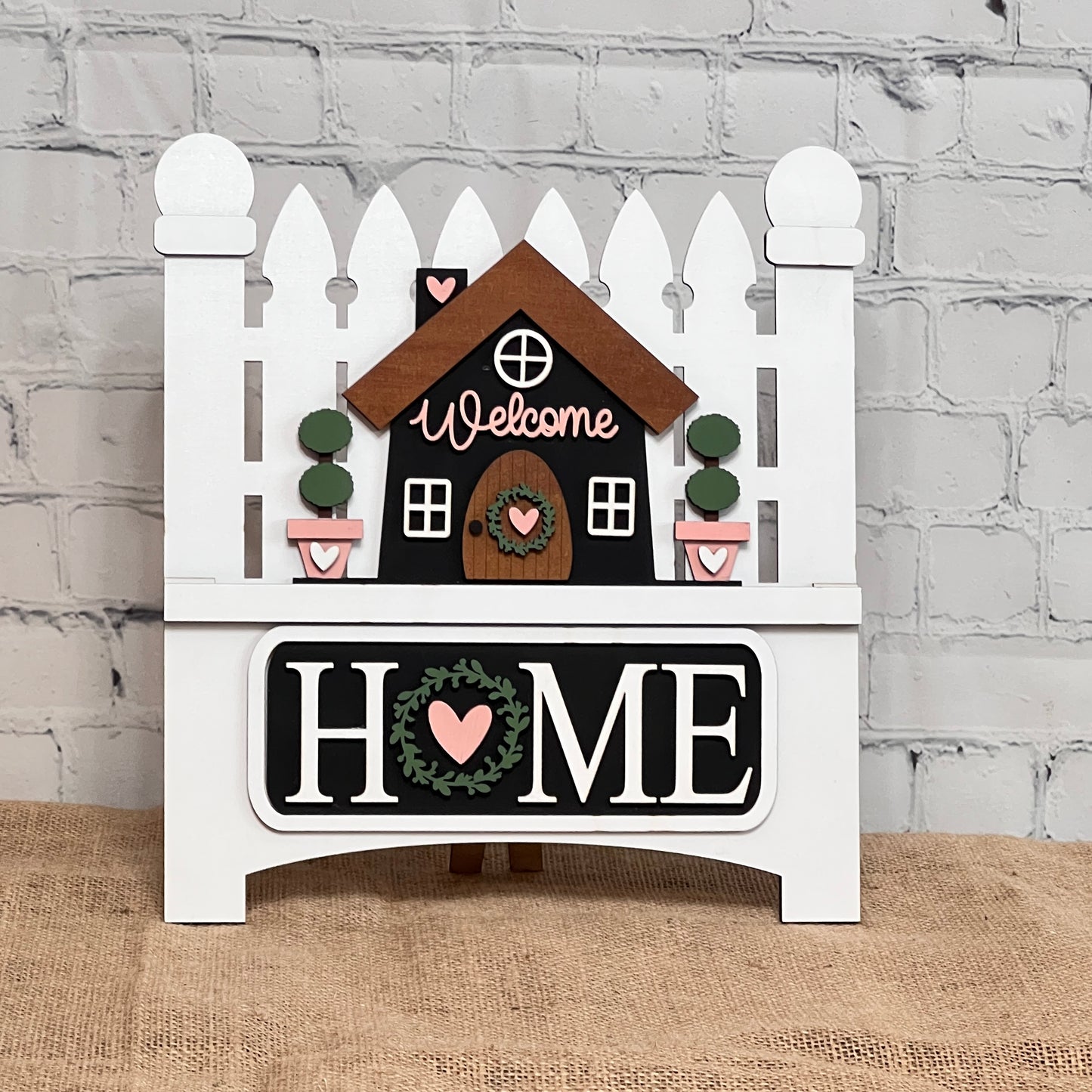 Janet's Craft Corner offers a DIY kit featuring a hand-painted picket fence sign with a house and "Welcome" text. "HOME" is bold with heart and wreath motifs, perfect for adding charm to brick walls or burlap surfaces, catering to home décor enthusiasts.