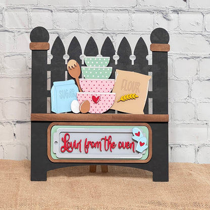 Janet's Craft Corner's Picket Fence with Insert kit features a kitchen sign with stacked bowls, eggs, and "sugar"/"flour" boxes on a shelf. Below it reads "Love from the oven," enhanced by heart accents on a white brick wall backdrop.