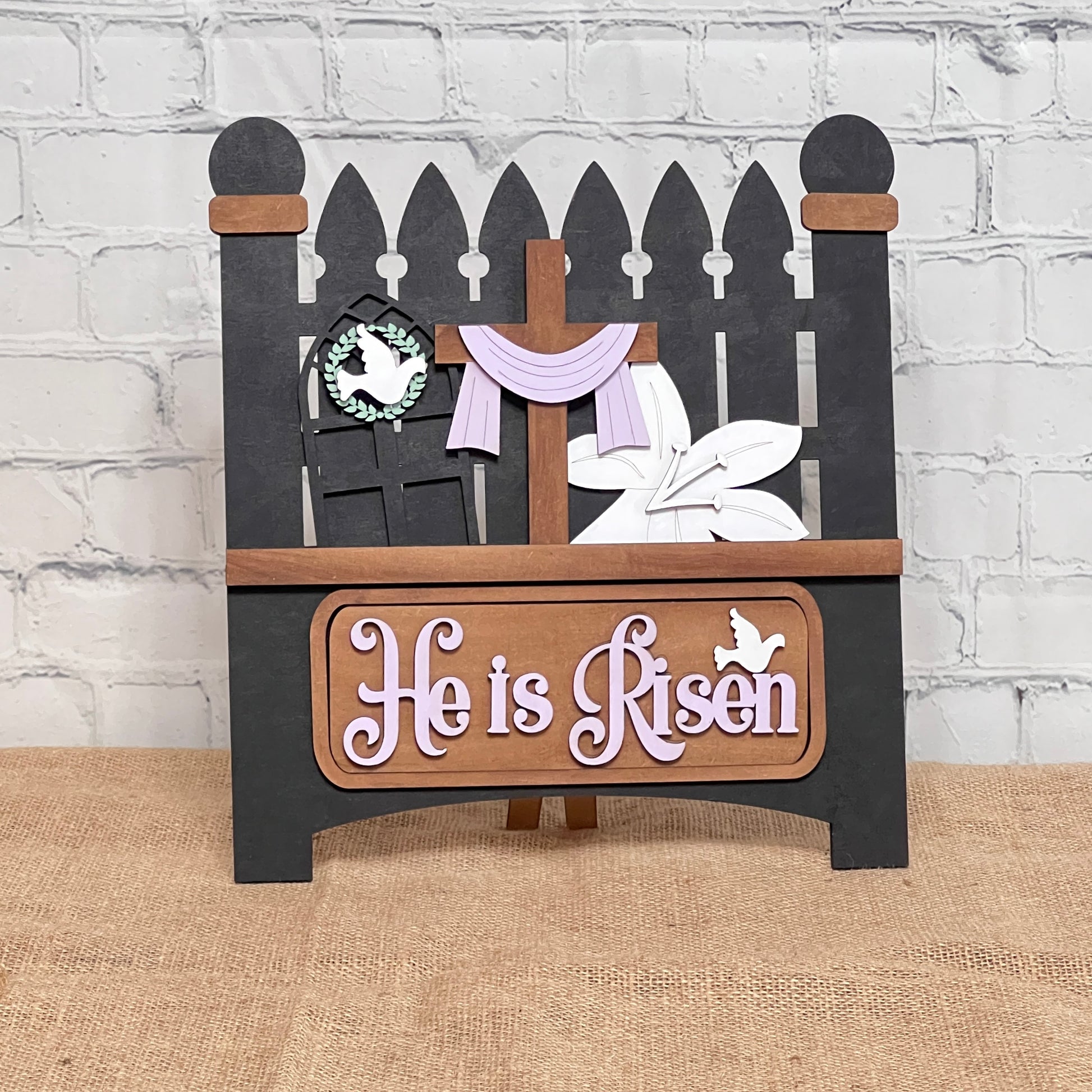 The Picket Fence with Insert from Janet's Craft Corner features a black fence with a hand-painted cross, draped white and purple cloth, a small wreath, white lilies, "He is Risen" text, and a white bird accent—perfect for adding charm to your home décor.