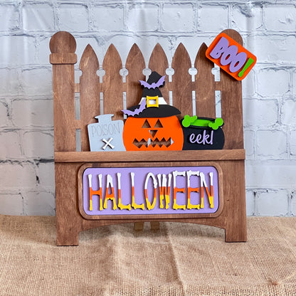 This delightful Halloween decor from Janet's Craft Corner features a wooden fence backdrop with interchangeable inserts: a pumpkin, witch's hat, "Poison" bottle, and bubbling cauldron. A cheerful "BOO!" sign hangs above, while vibrant letters spell "HALLOWEEN" at the base.