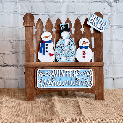 The Winter Wonderland Interchangeable Insert DIY Craft Kit by Janet's Craft Corner features a charming decorative piece depicting a garden fence with three smiling snowmen. The area below the snowmen is adorned with the words "Winter Wonderland" and on the top of the right post is "BRRR" in white and blue, all  against a white brick wall and placed on a burlap surface. It's perfect for interchangeable home decor!