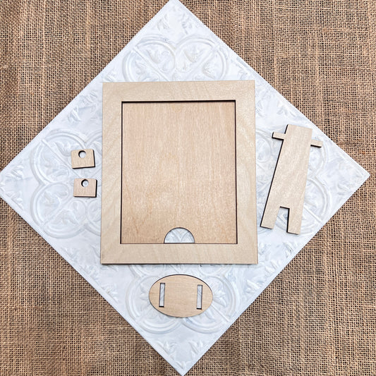 Pieces from Janet's Craft Corner's Simple Farmhouse Frame Shelf Sitter DIY Kit are arranged on a white embossed tile over burlap. Shown: interchangeable sign frame, stand, and connectors.