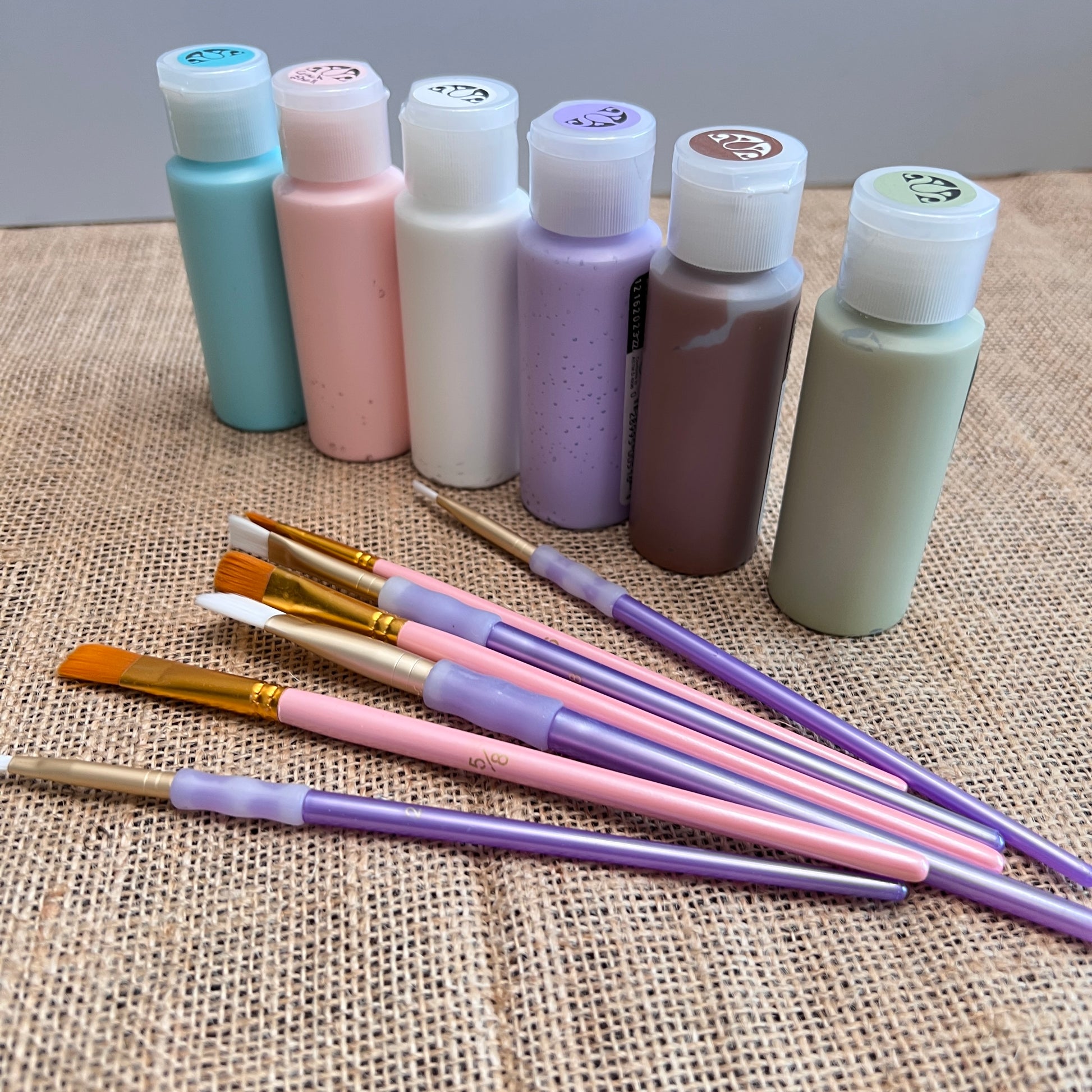 Arranged on burlap, six pastel paint bottles and eight pink and purple-handled brushes form a charming DIY kit. Perfect for crafting with Janet's Craft Corner's Bunny Table Top Pallet, a seasonal home decor masterpiece!.