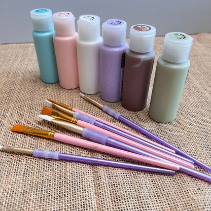 Arranged on burlap, six pastel paint bottles and eight pink and purple-handled brushes form a charming DIY kit. Perfect for crafting with Janet's Craft Corner's Bunny Table Top Pallet, a seasonal home decor masterpiece!.